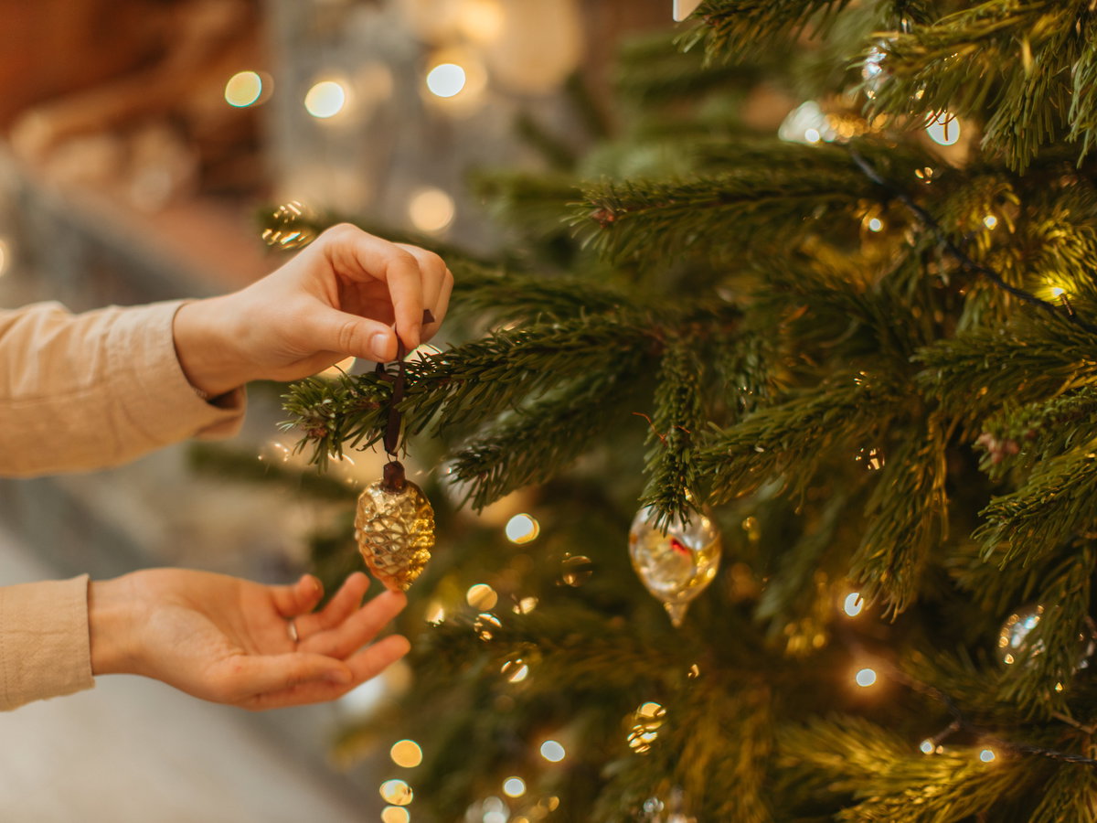 Frau hängt goldenen Christbaumschmuck an einen Tannenbaum im Wohnzimmer