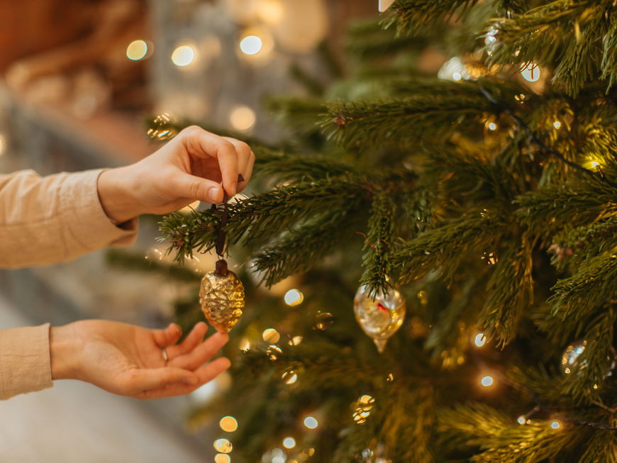 Frau hängt goldenen Christbaumschmuck an einen Tannenbaum im Wohnzimmer