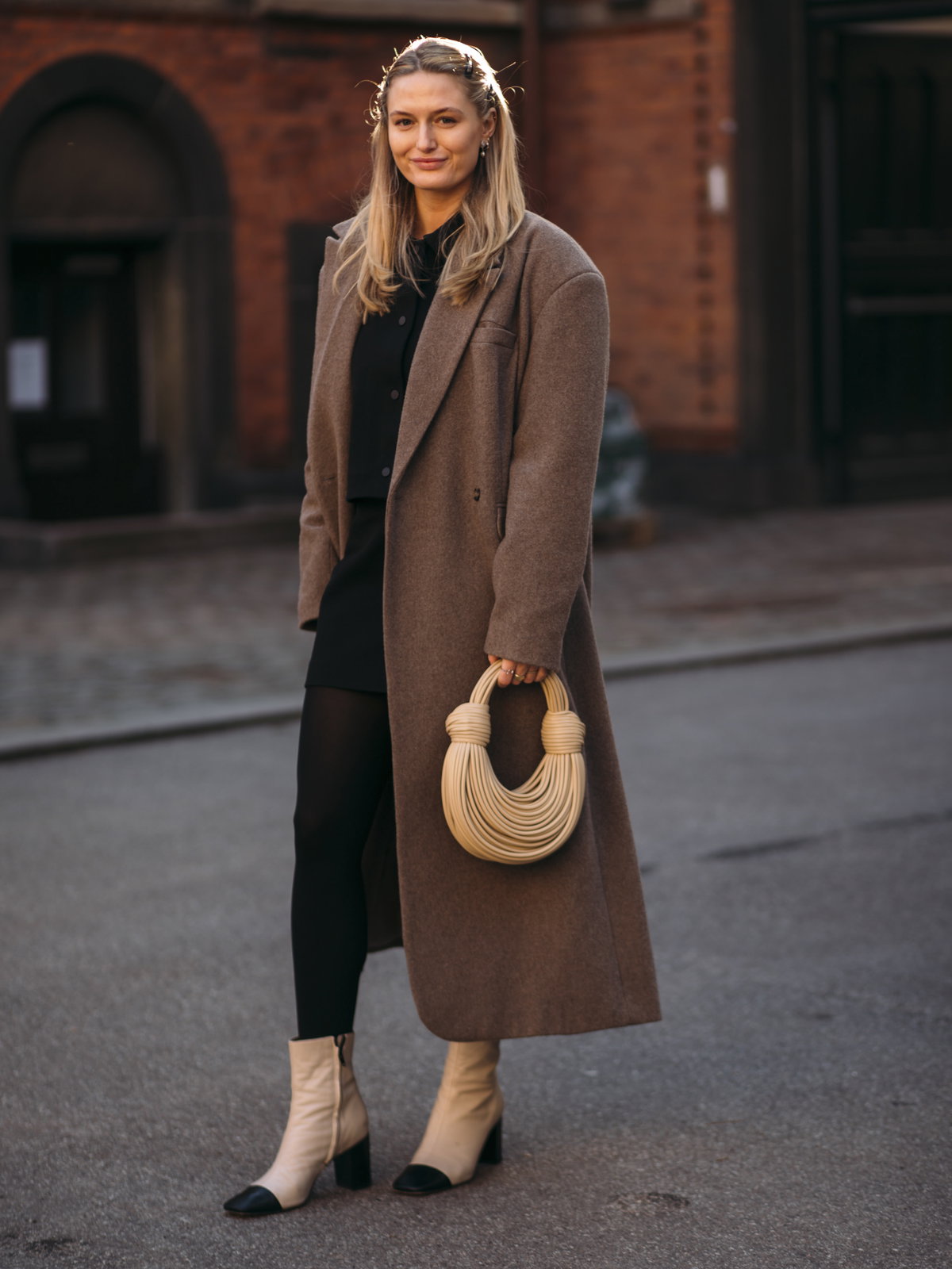 Frau trägt auf Kopenhagen Fashion Week Ankle Boots schwarzen Rock braunen Mantel und cremefarbene Tasche Ankle Boots kombinieren