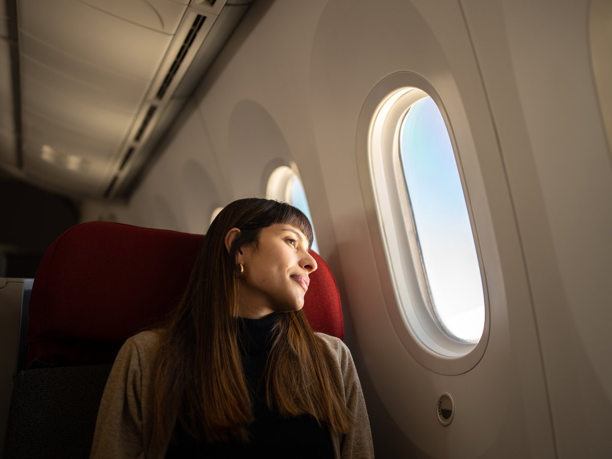 Frau mit langen, braunen Haaren sitzt im Flugzeug und schaut aus dem Fenster