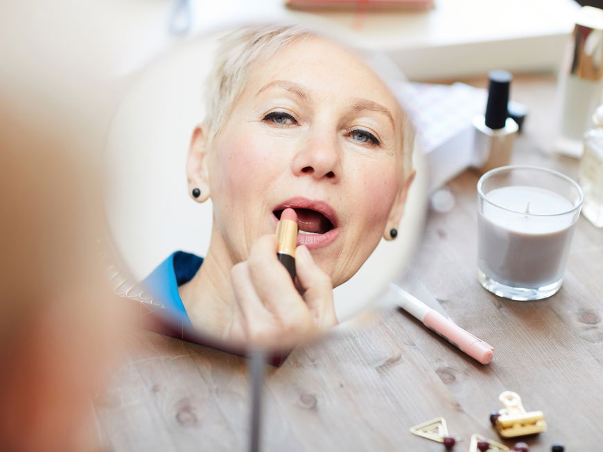 Frau mit grauen Haaren trägt vor dem Spiegel Lippenstift auf.