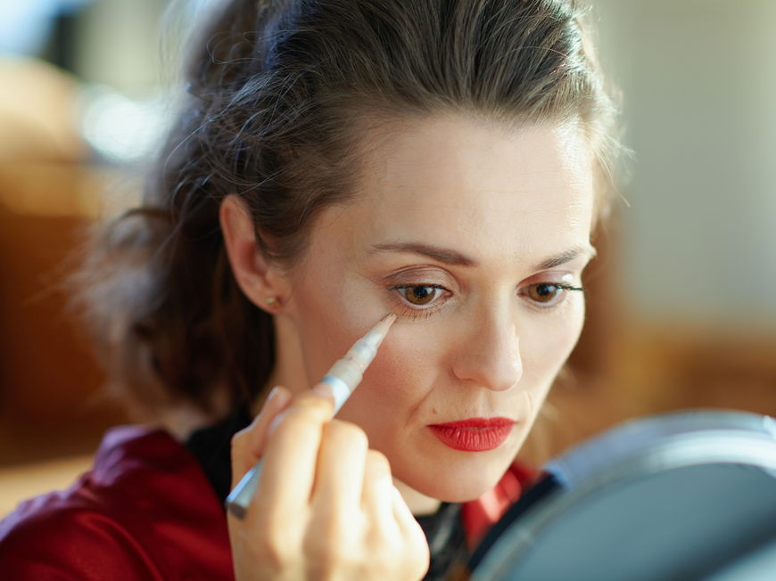 Frau sitzt vor dem Spiegel und deckt ihre Augenringe mit Concealer ab.
