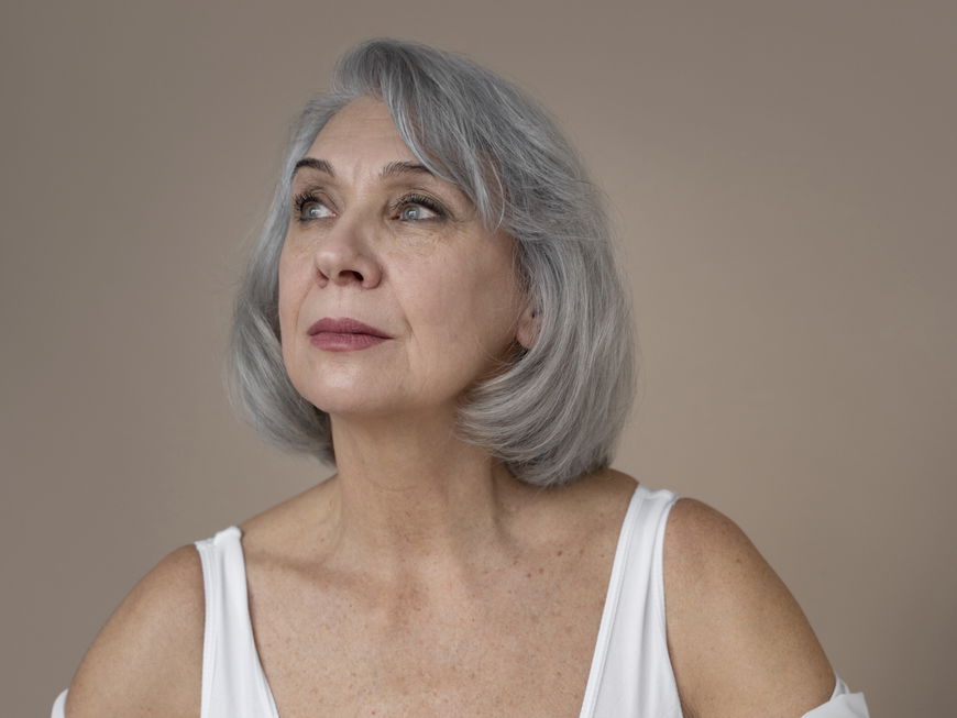 Frau mit grauen kurzen Haaren trägt eine weiße Bluse und Lippenstift.