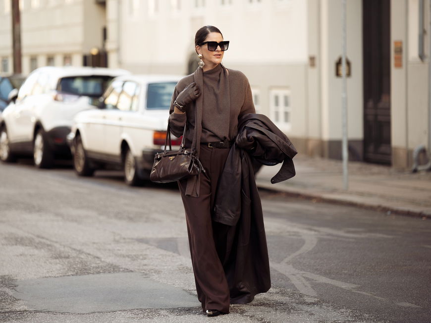 Frau trägt ein komplett braunes Outfit mit Hose, Pullover und Tasche und einer Sonnenbrille.