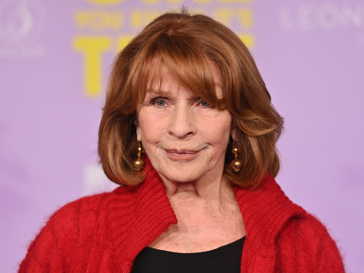 Senta Berger mit goldenen Ohrringen und einer roten Strickjacke.