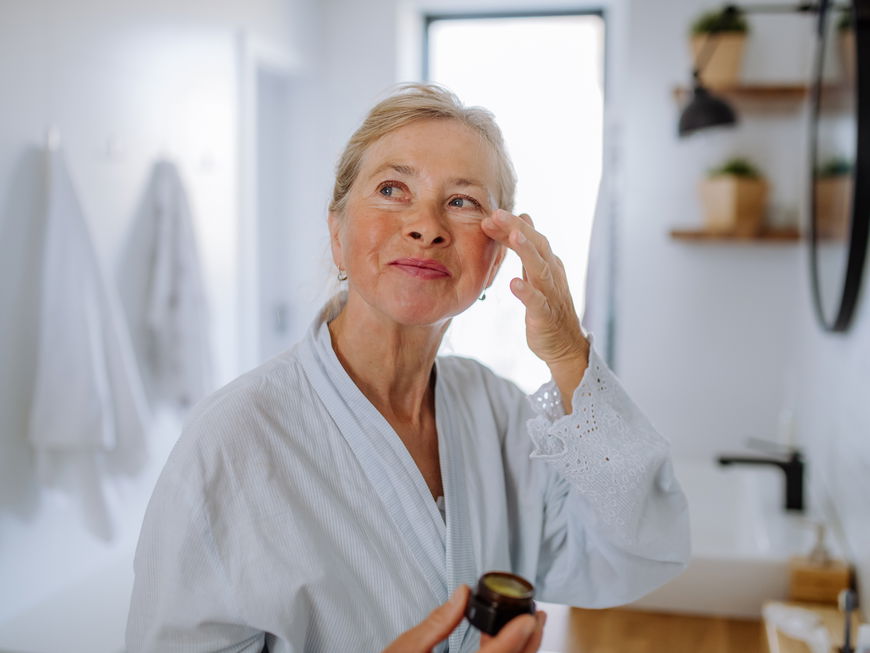 Ältere Frau trägt im Bademantel eine Creme unter ihren Augen auf.