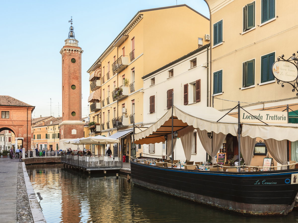 Trattorie und Häuser mit Blick auf den Kanal, der durch Comacchio fließt. Im Hintergrund plaudern Einheimische