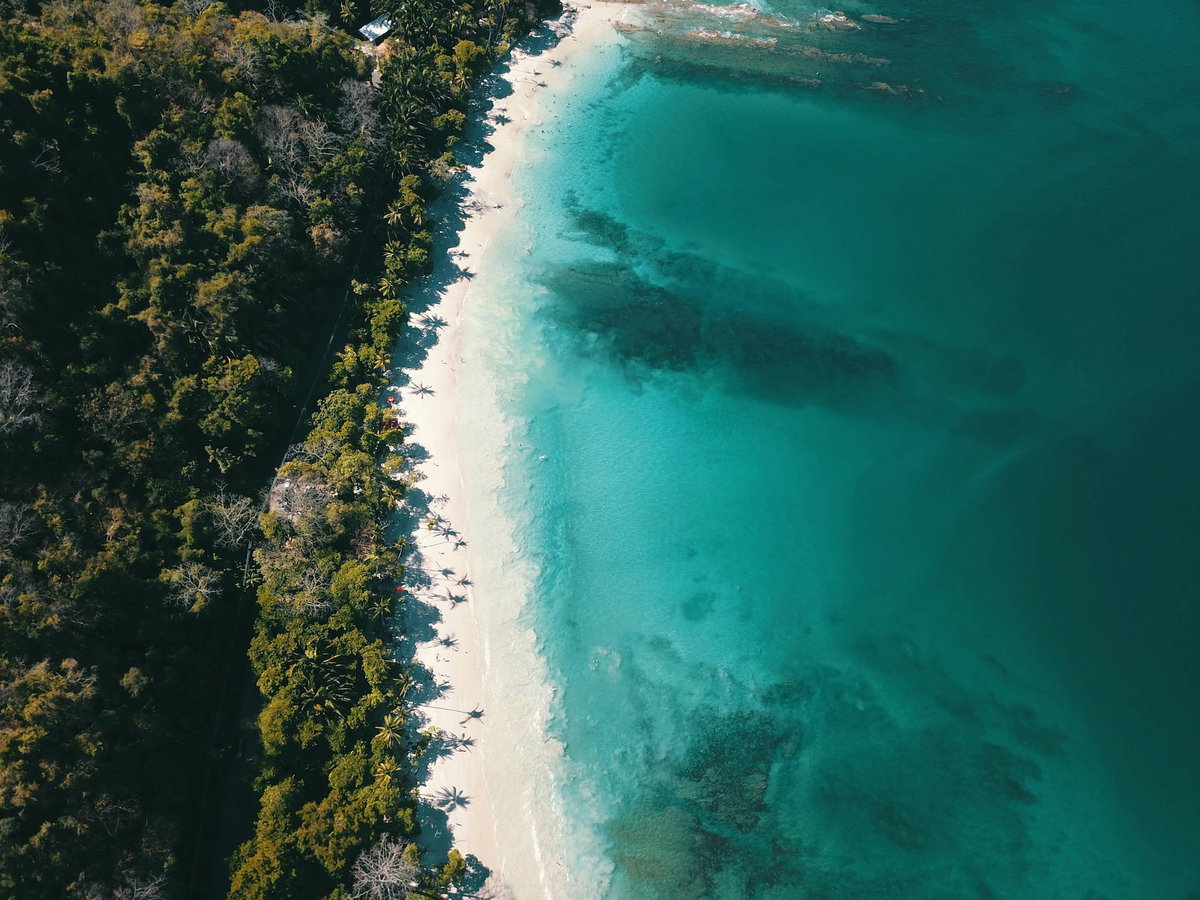 Blick auf den Strand, türkisblaues Wasser und grüne Landschaft aus der Vogelperspektive.