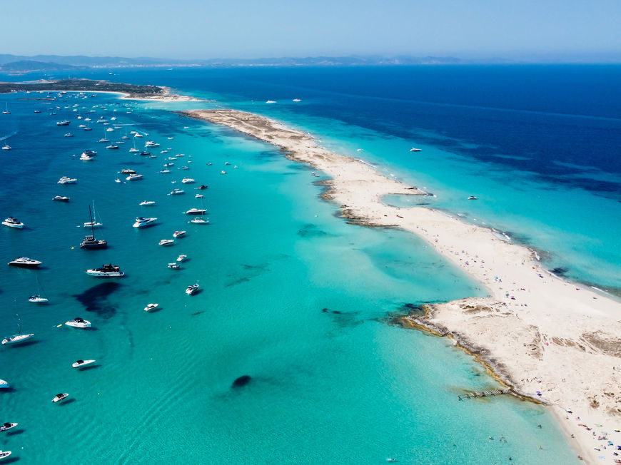 Ein langer Sandstrand mit türkisblauem Meer und Yachten darin.