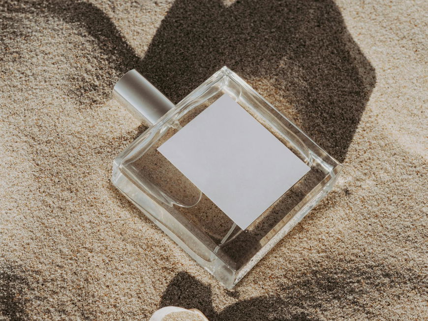 Ein Flakon liegt im Sand mit einer Muschel daneben.