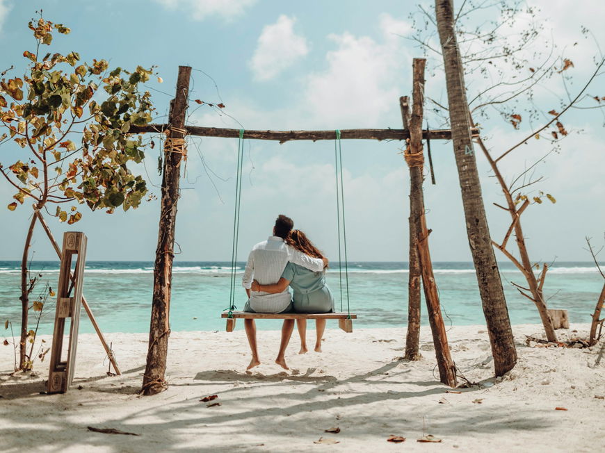 Pärchen von hinten sitzt Arm in Arm auf einer Schaukel am Strand.