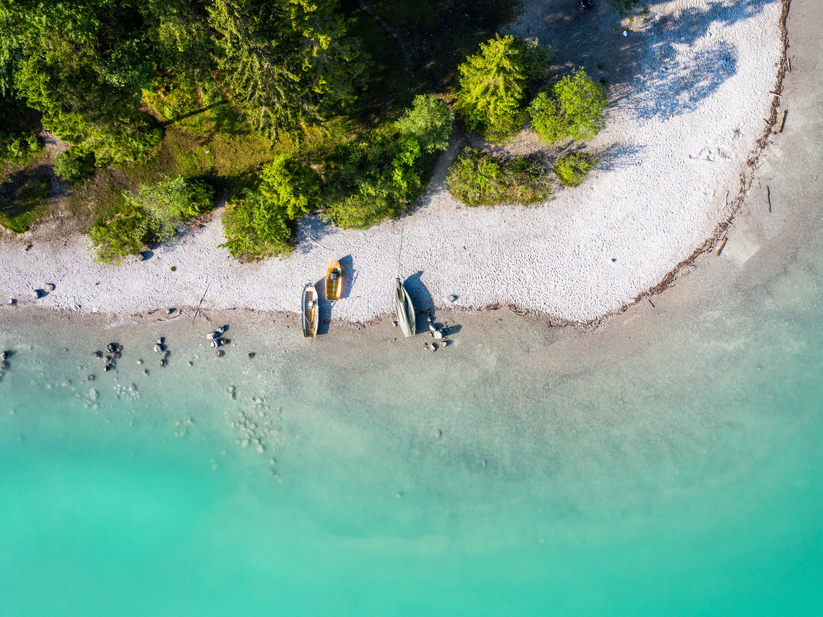 Ein Strand mit türkisblauem Wasser, Fischerbooten und Nadelbäumen von oben.