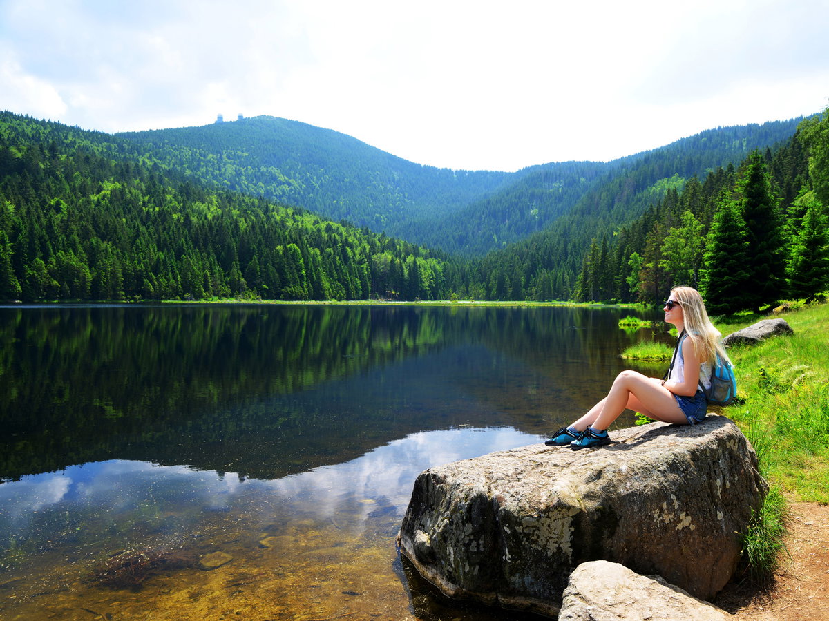 Frau sitzt auf einem Stein an einem See mit Wäldern im Hintergrund.