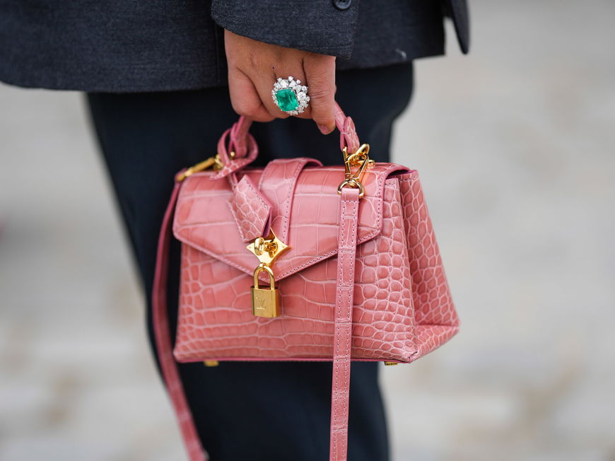 Eine kleine rosane Tasche wird von einer Hand mit einem Smaragd-Ring gehalten.
