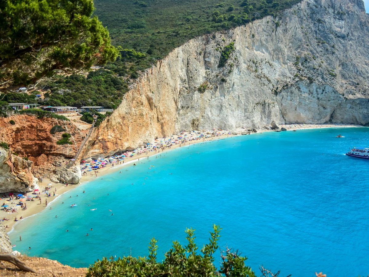 Ein Blick auf einen schmalen Strand mit einem großen Hand dahinter. An dem Strand sind einige Menschen und Sonnenschirme.