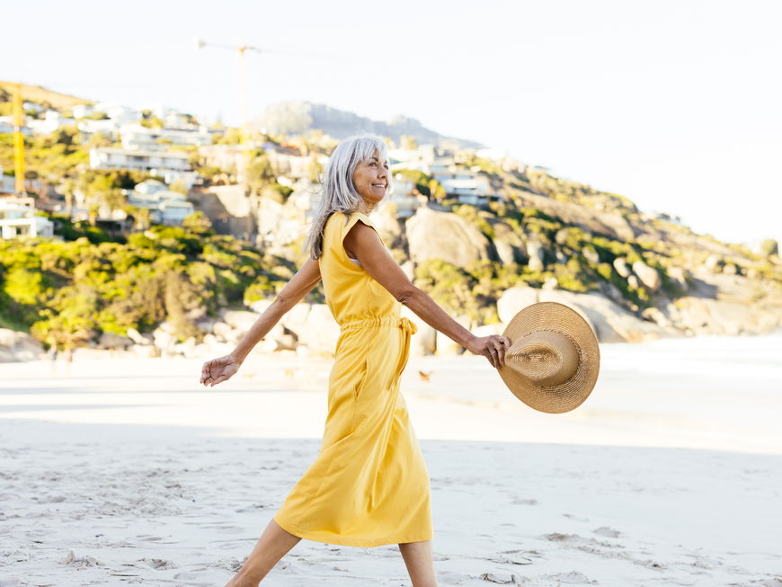 Frau mit grauen Haaren trägt ein langes, gelbes Kleid und spaziert am Strand mit einem Sonnenhut.