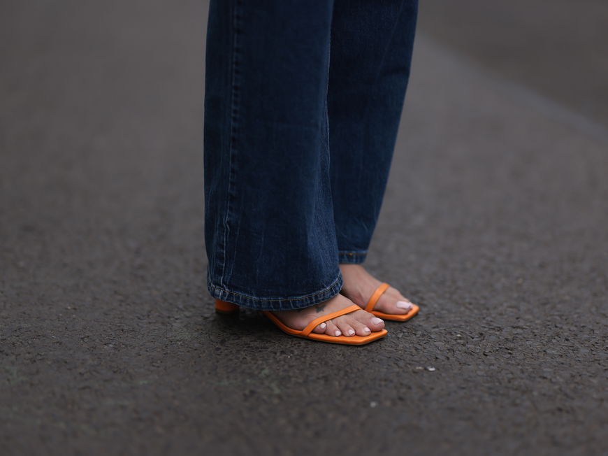 Nackte Füße in orangenen Sandalen mit hellem Nagellack und einer langen, dunklen Jeans.