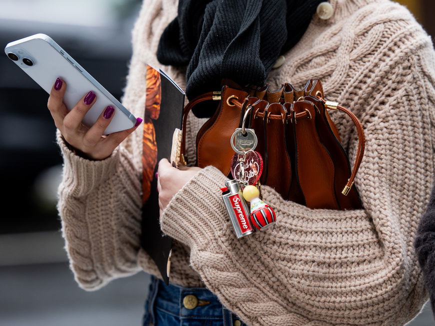 Frau mit einem Cardigan hält ihre Tasche unterm Arm und ein Handy in der Hand. An der Tasche ist ein Schlüsselanhänger.