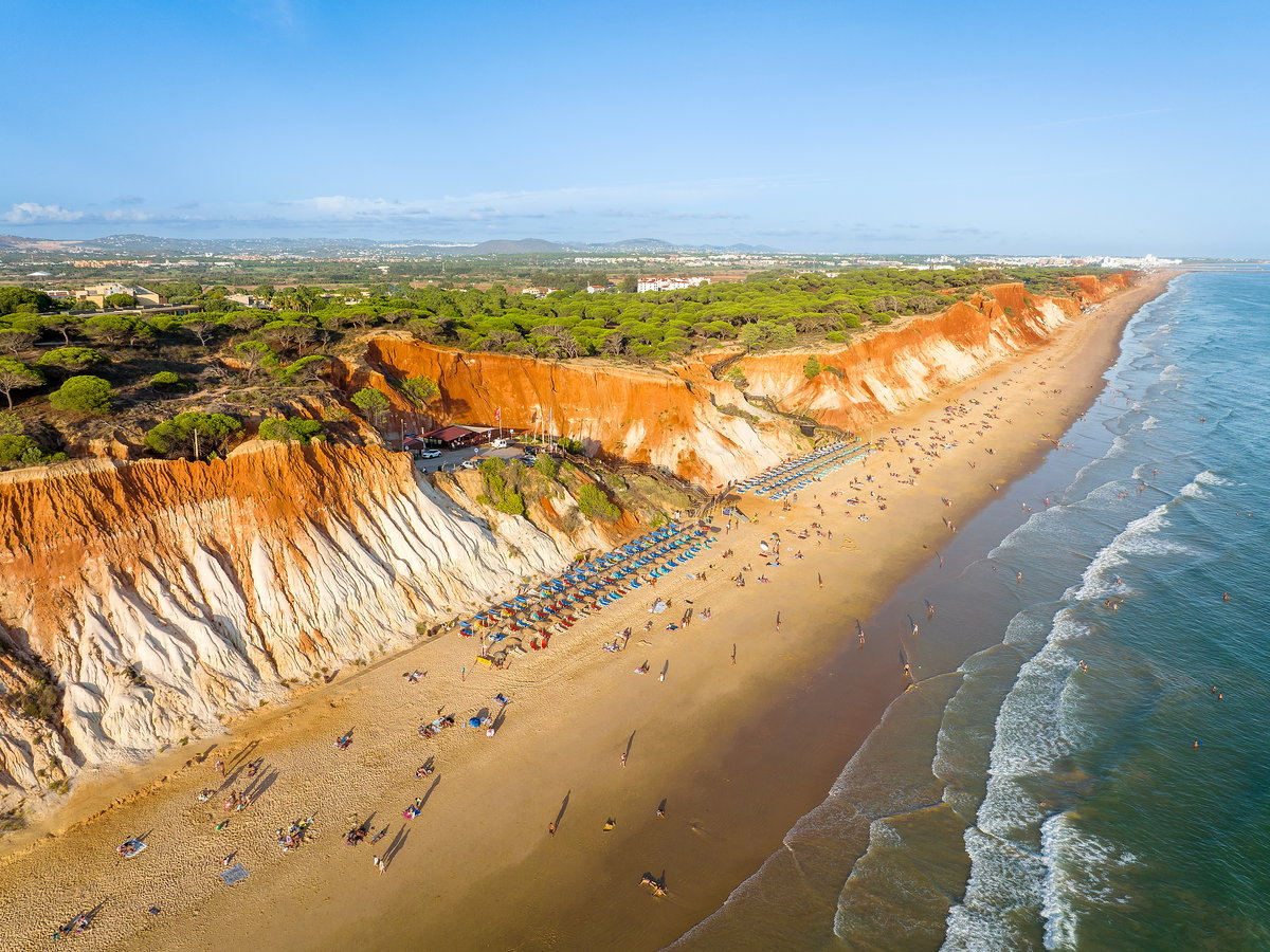 Ein Strand mit rötlichen Felsen, Bäumen darauf und Sonnenschirmen und -liegen am Strand.