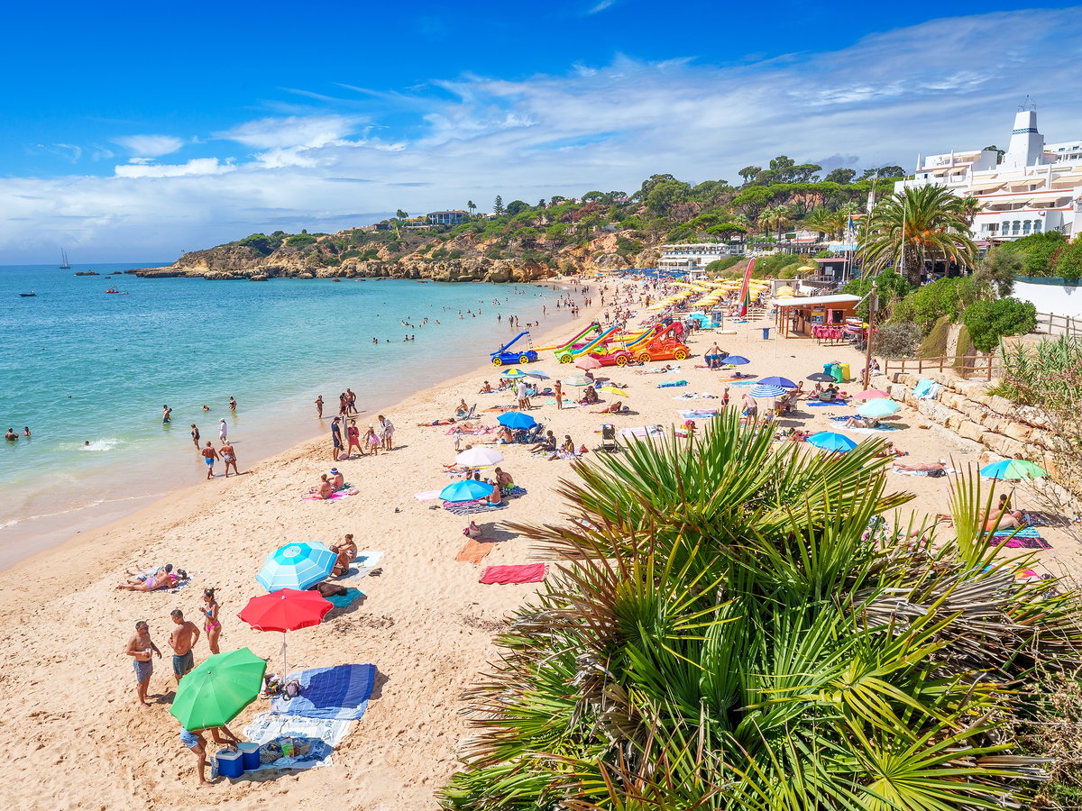 Strand mit vielen Urlaubern, Sonnenschirmen, Palmen und Tretbooten am Strand.