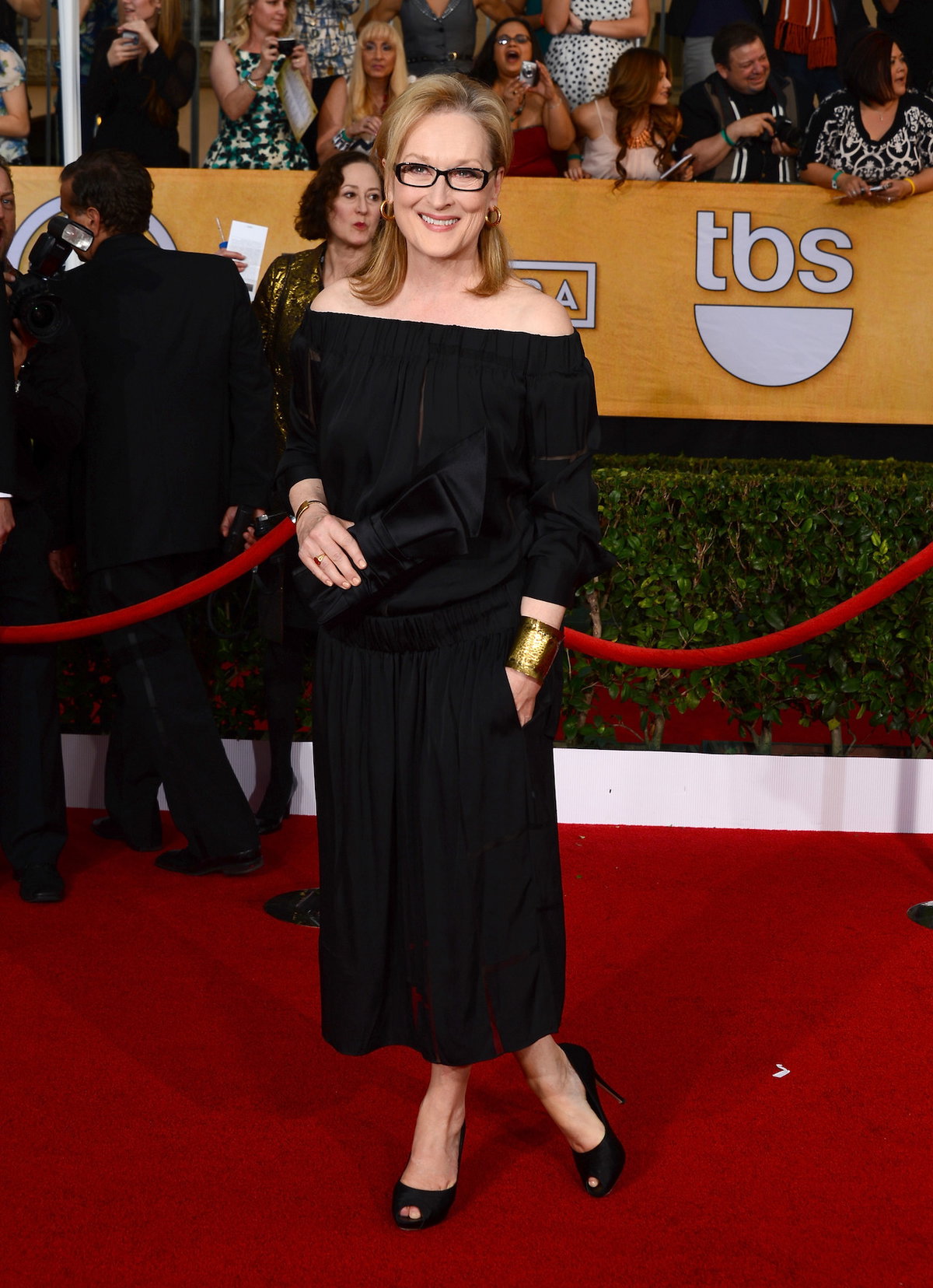 Meryl Streep mit einem schwarzen, langen Kleid und einer schmalen Brille. Im Hintergrund sind Menschen.