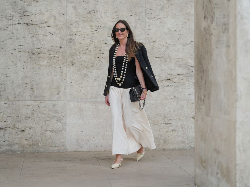 Frau trägt einen weißen Maxi-Rock im Boho-Stil mit einem schwarzen Top und Blazer und zwei großen Perlenketten.