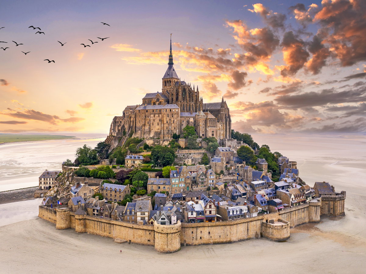 Ein Blick auf den Mont Saint Michel bei Sonnenaufgang mit Wolken und Vögeln am Himmel.