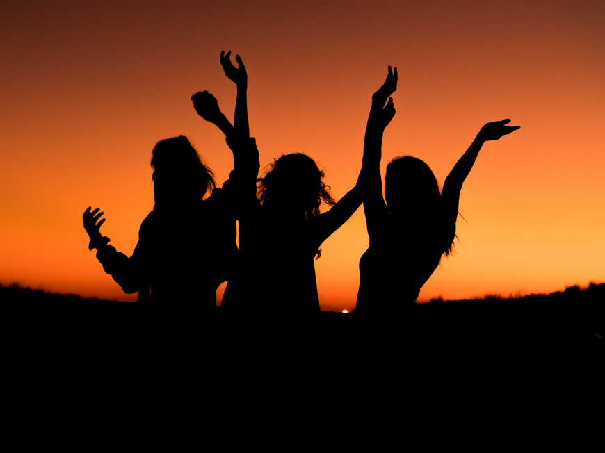 Drei Frauen tanzen ausgelassen vor dem Sonnenuntergang.