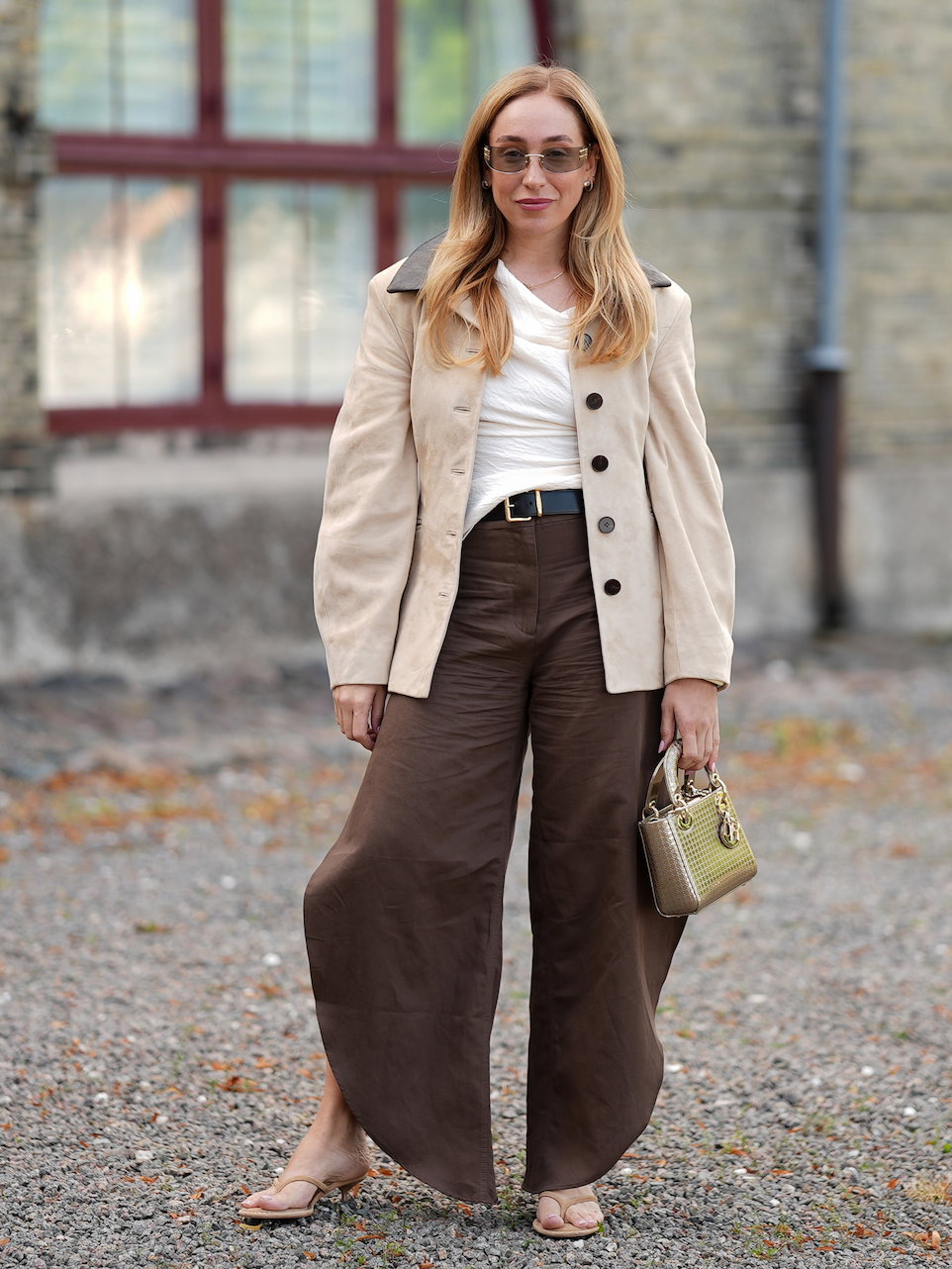 Frau mit blonden Haaren, Sonnenbrille, hellem Shirt, dunkelbrauner Hose und beigem Blazer.