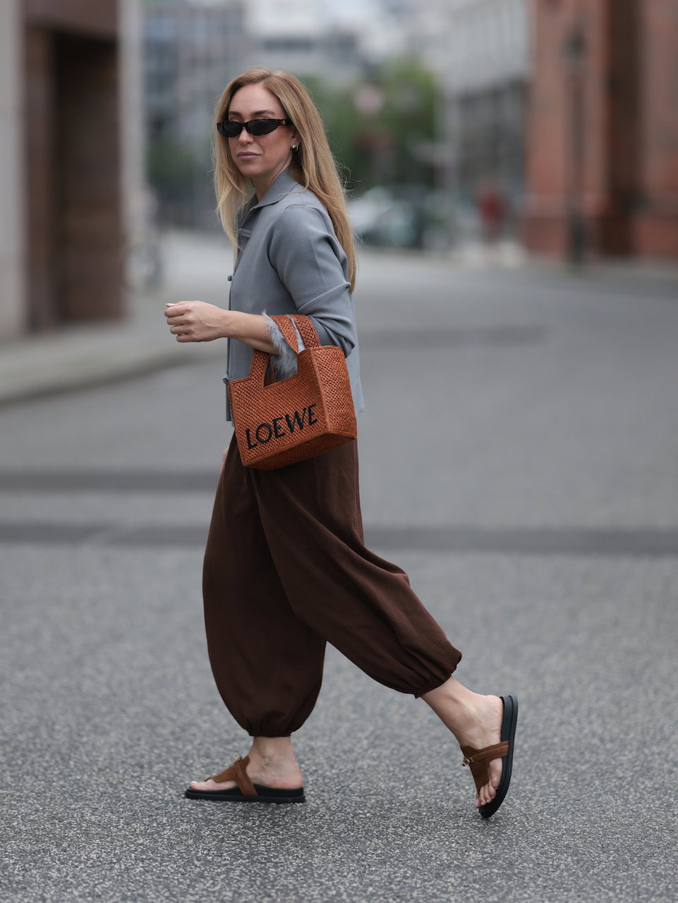 Frau mit blonden Haaren, Sonnenbrille, grauem Blazer und dunkelbrauner Hose mit FlipFlops.