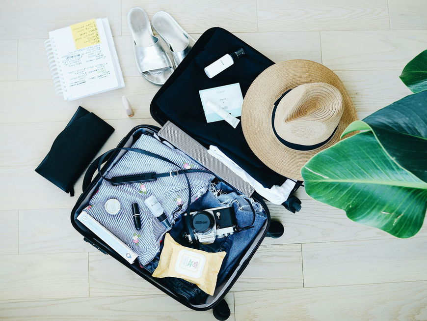 Koffer auf dem Boden mit einer Kamera, Kleidung, Beauty-Produkten, Schuhen und einem Hut.