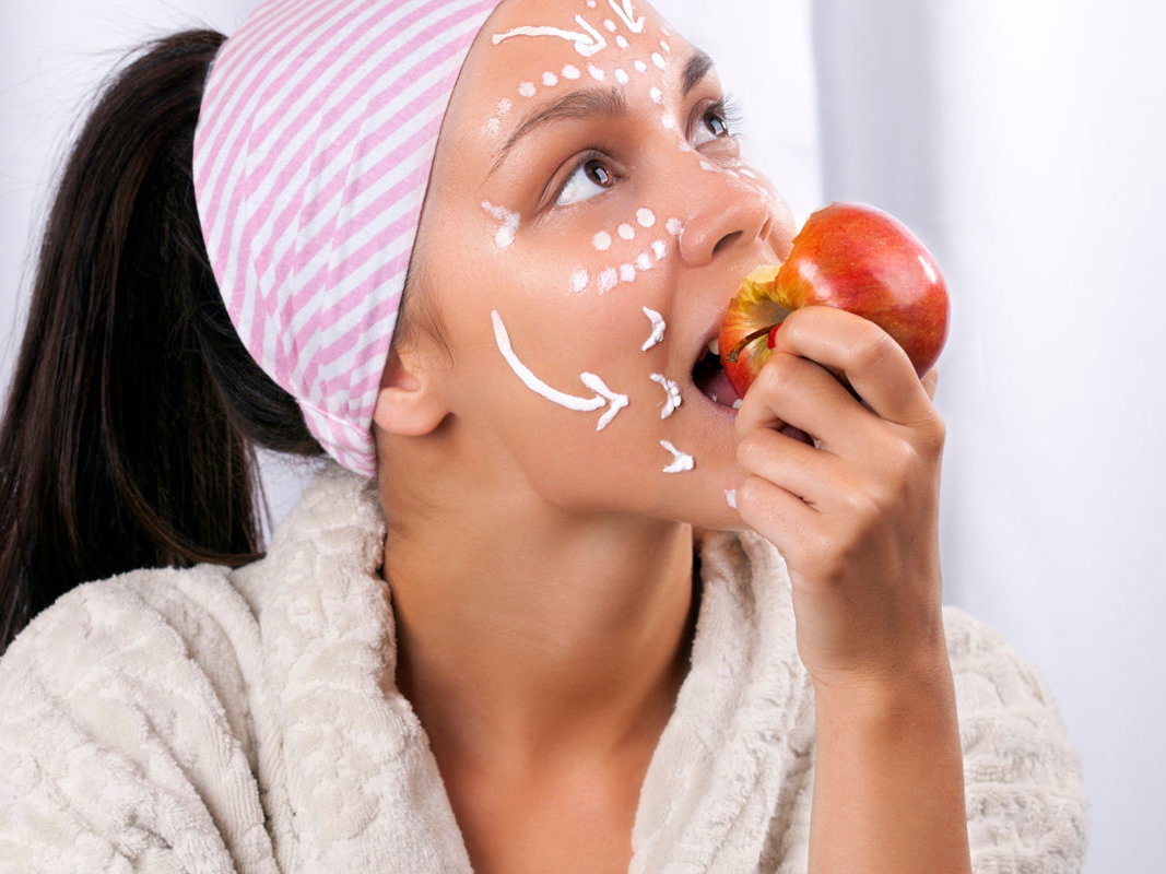 Frau mit Pflegecreme im Gesicht isst Apfel